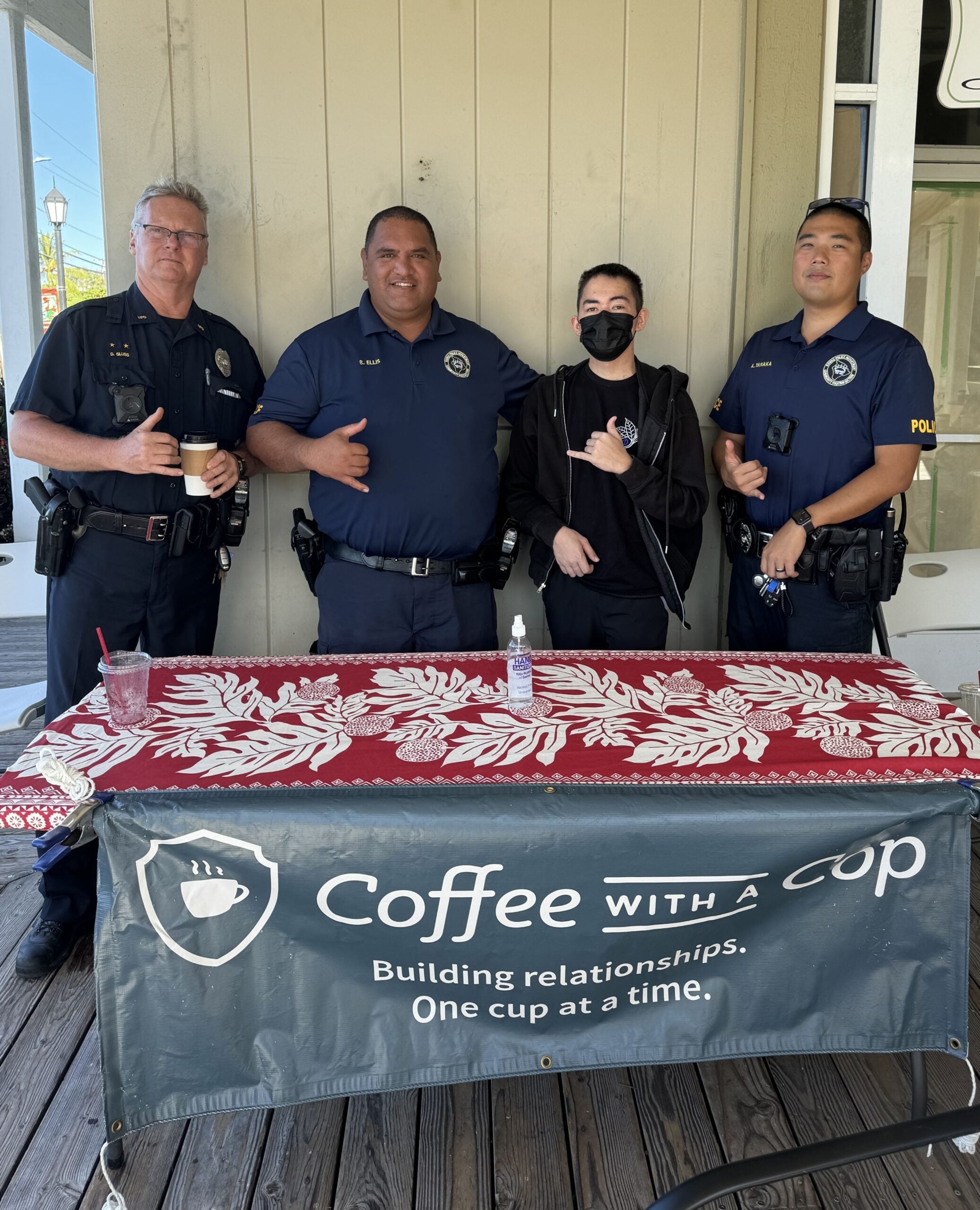 Three police officers standing with young man by table at coffee shop with banner that says Coffee With a Cop.