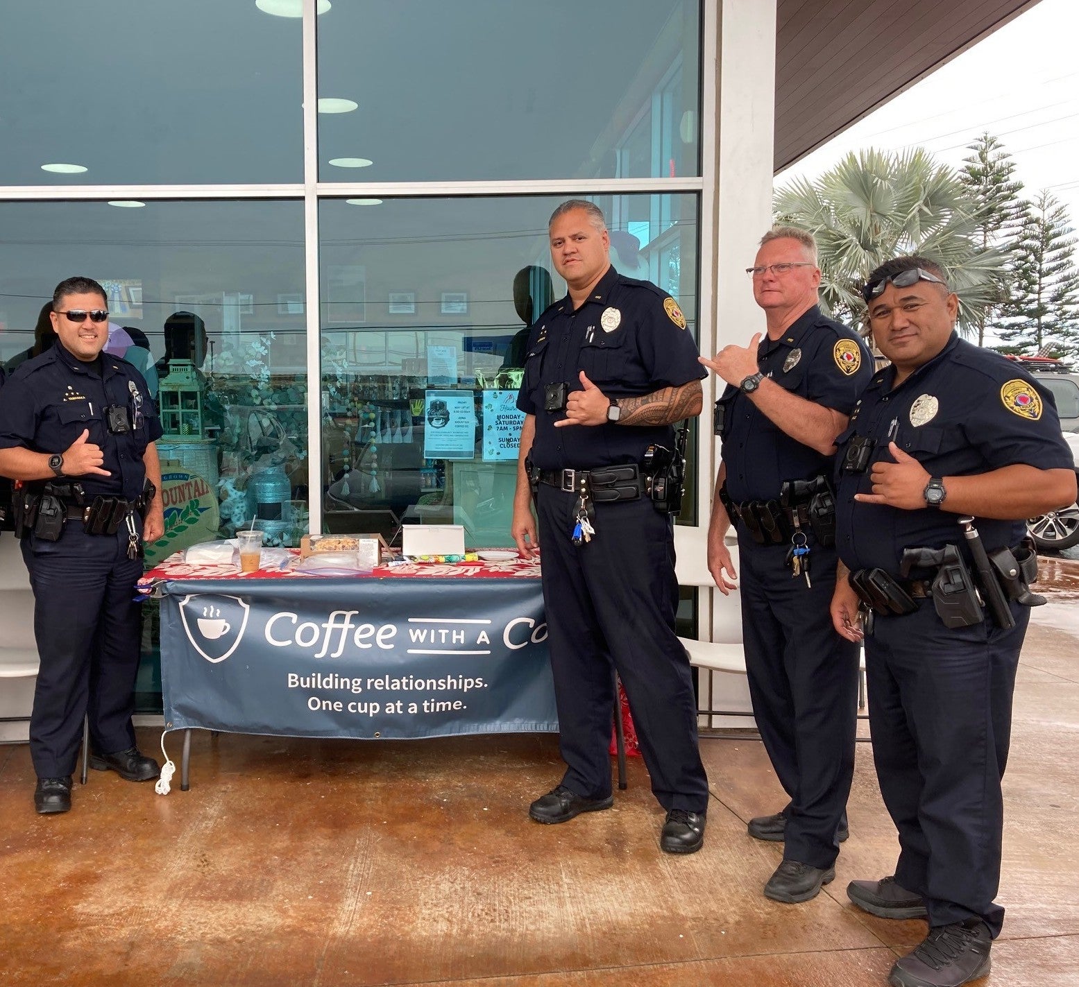 4 police officers standing by table at coffee shop with banner that says Coffee With a Cop.