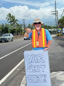 Man waving on sidewalk holding a sign that says car seat check. 