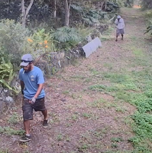 Man in foreground with blue shirt, black shorts with his head down smoking a cigarette. Man in background with grey shirt and dark grey shorts. Can not see his face due to head covering.